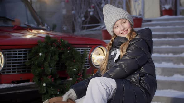 Portrait Smiling Girl on Red Car with Christmas Tree Branch Decoration Background in Winter Day