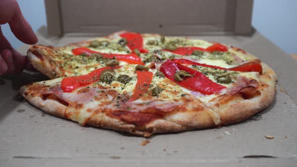 Hands taking slices of a delicious pizza with ham, cheese, red peppers, and olives.