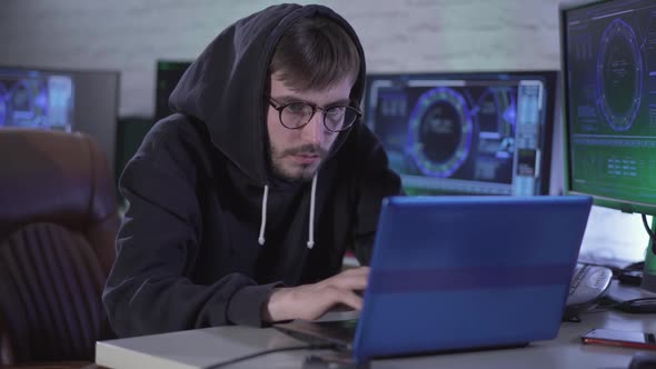 Serious Bearded Man in Eyeglasses and Hood Typing on Laptop Keyboard and Looking at Computer Screen