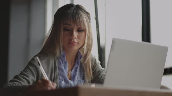 Asian Woman Working on Her Laptop and Writing in His Notebook Sitting at a Table