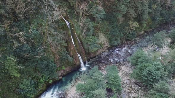 A small waterfall flows into the mountain river. Around the mountains and green trees. Aerial view.