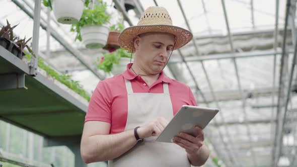 Portrait of Focused Man Standing in Greenhouse and Typing on Tablet. Front View of Confident