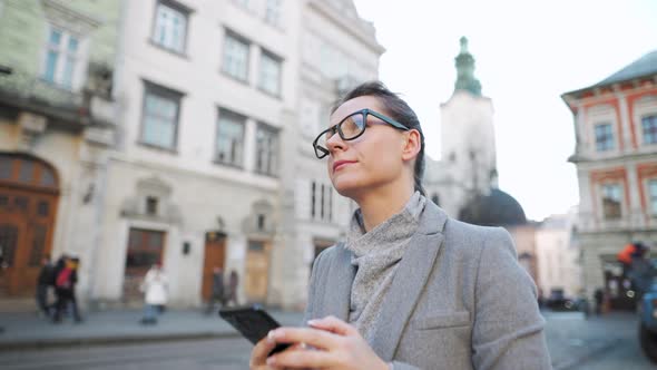 Woman with Glasses Wearing a Coat Walking Down an Old Street and Using Smartphone