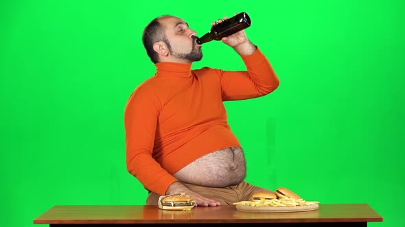 Overweight Caucasian Man Sits at a Table with Junk Food, Drinks Beer and Lets Out a Good Burp, Green