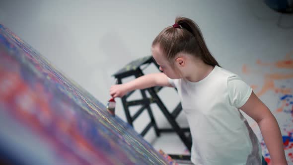 Kid Girl with Down Syndrome Draws with a Brush on a Large Canvas in a White Room Kid Girl with