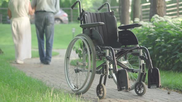 Wheelchair Standing on Park Alley in Sunlight with Blurred Senior Caucasian Couple Walking Away at