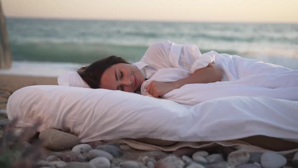 Happy Young Smiling Woman Sleeping in White Soft Bed with Turquoise Sea Waves Splashing with Foam on