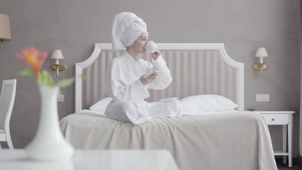 Portrait of Young Caucasian Woman in White Bathrobe and Hair Towel Drinking Coffee and Using Social