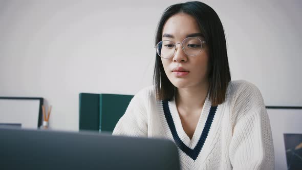 Young Chinese Female Entrepreneur in Casual Outfit is Looking Thoughtful While Working Sitting at a