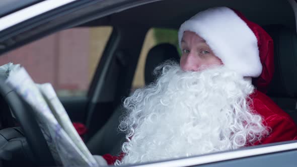 Confused Bearded Santa Clause Sitting in Car on Driver's Seat Examining Paper Map