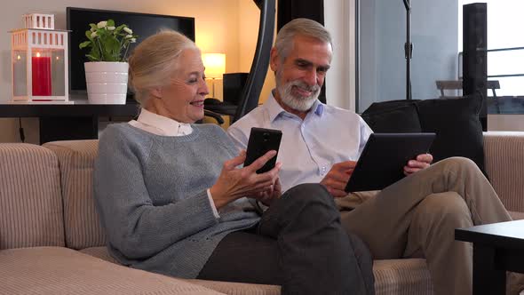 An Elderly Couple Sits on A Couch in An Apartment and Works on A Smartphone and A Tablet