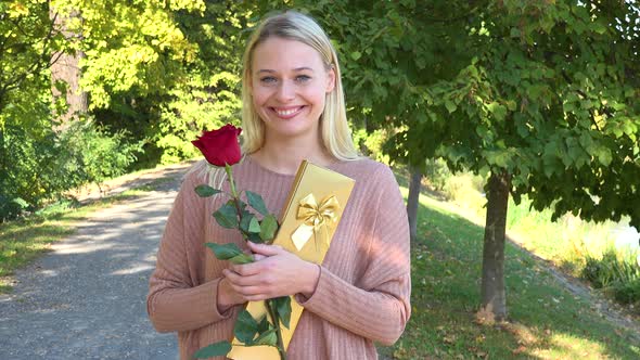 A Young Beautiful Woman Holds a Rose and a Present and Smiles Happily at the Camera in a Park