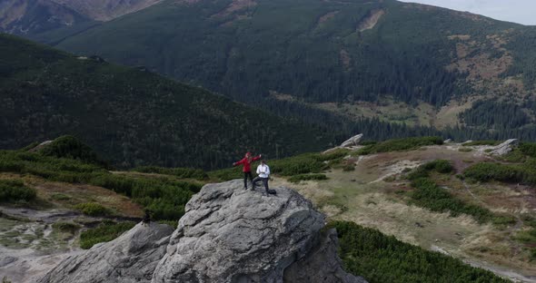 a girl waving a hand a guy controls a drone a black dog stands on a big rock in the mountains