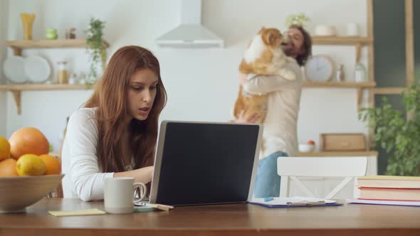 Attractive Woman Trying To Work Remotely While Her Husband Holding in the Arms a Dog and Dance.