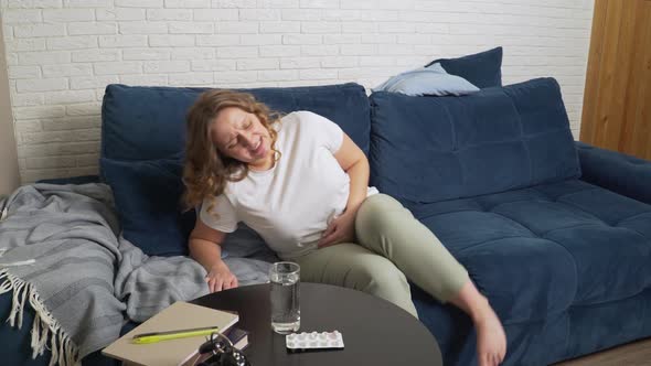 Young Woman Lies Down on the Blue Sofa Massages Her Stomach Due to Severe Pain and Drinks Water