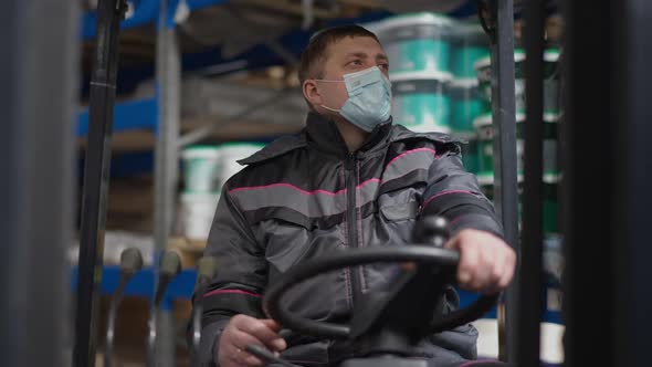 Front View of Serious Man in Uniform and Covid Face Mask Sitting in Warehouse Autoloader Turning