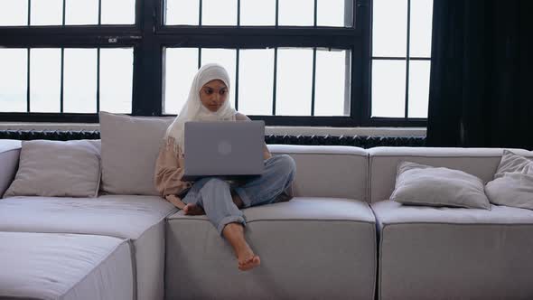 Young Muslim Girl Sits on the Couch Looking at the Laptop in Bright Beige Studio