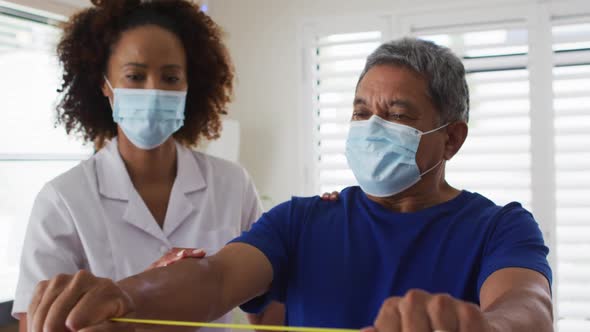 Mixed race female physiotherapist wearing mask helping senior exercise using exercise band