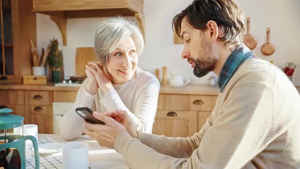 Young Man Teaching His Elderly Mother to Use Mobile Phone Explaining Online Communication Rules