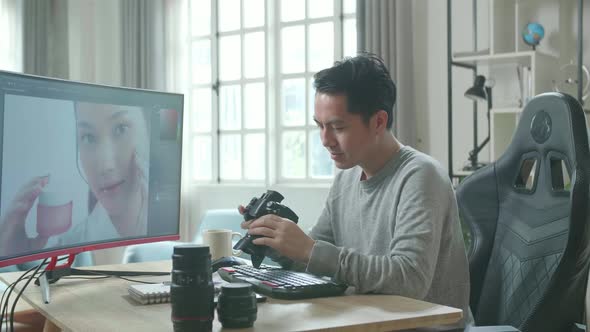 Asian Cameraman Holding And Looking At Camera While Using Desktop Computer For Editing At Home Photo