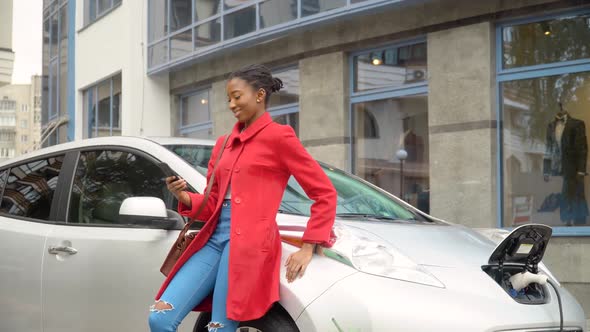 African American Girl Using Smartphone and Leans on Electric Car That Is Charging. Plug Charging an