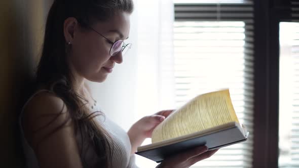 Girl with glasses reads a book near the window with shutters, back light