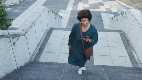 Plus size biracial woman walking on stairs with smartphone in city