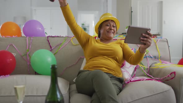 Happy african american senior woman using party blower making new year's eve tablet video call