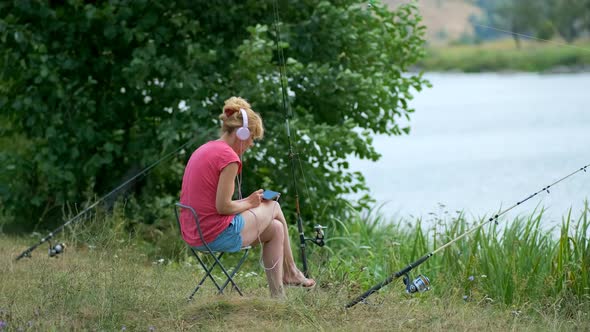 A young girl fishing on a spinning rod sitting on the Bank of the river in headphones.