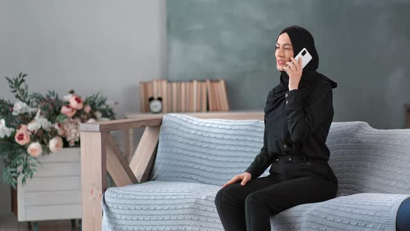 Happy Young Muslim Woman in Traditional Black Hijab Talking Use Smartphone Sitting on Couch