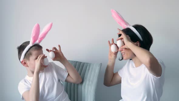 Playful boy and woman with bunny ears keeping white eggs near eyes on white background. Happy child