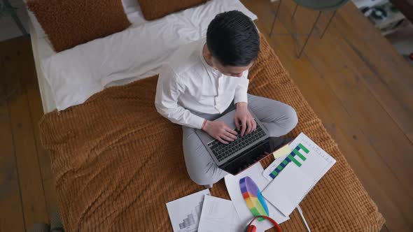 Top View of Concentrated Asian Young Man Typing on Laptop Keyboard Sitting on Bed at Home Office