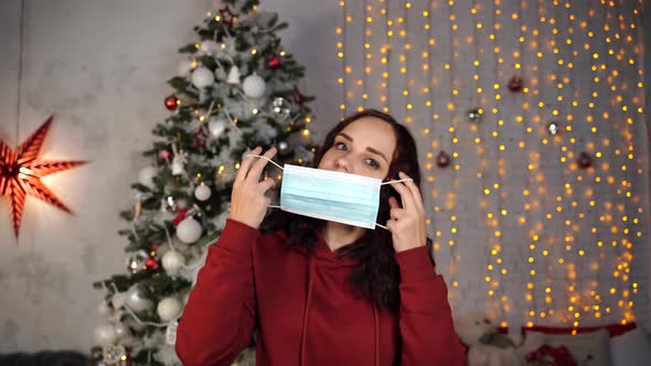 Young Woman Putting Medical Mask on Face on Background of Christmas Tree. Charming Brunette in