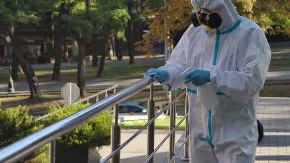 A Virologist in Protective Clothing Disinfects the Railing with an Antiseptic Spray and a Tissue