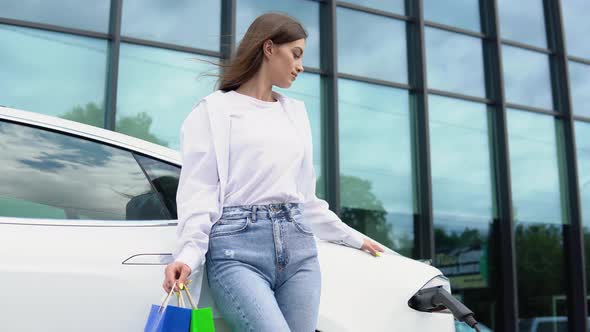 Young Girl After Buying Products with a Shopping Bag is Standing Near the Charging Electric Car