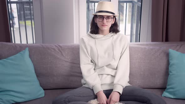 Girl in Glasses and a Hat Sits on the Sofa with Her Legs Crossed Window Background