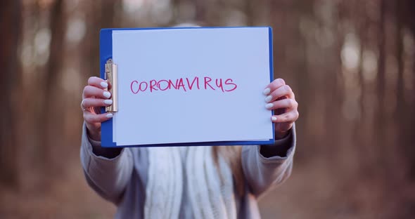 Woman with Protective Mask Holding Coronavirus Inscription in Hands in Forest alt