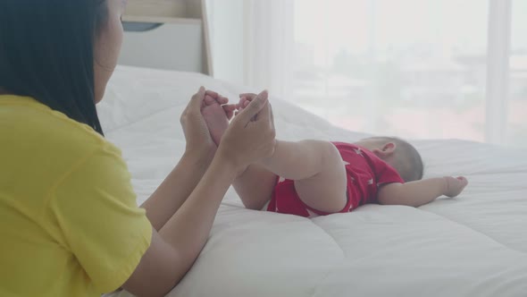 Young asian mother touch foot of little baby girl with tender on bed in the bedroom.