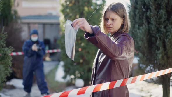 Young Female Investigator Examining Knife in Transparent Pack As Colleague Taking Photo of Crime