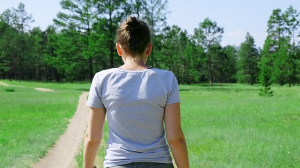 A Girl Walks Along a Country Road in a Green Meadow Surrounded By Forest