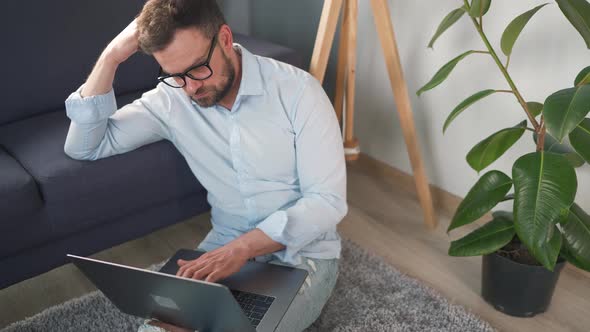 Man with Glasses Sitting on Carpet with Laptop and Smartphone and Working in Cozy Room