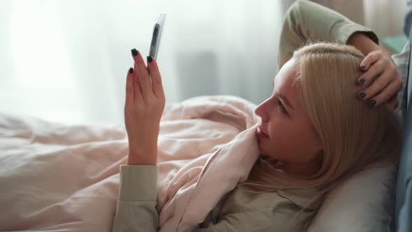 Concentrated blond woman hiding behind a blanket and looking at phone