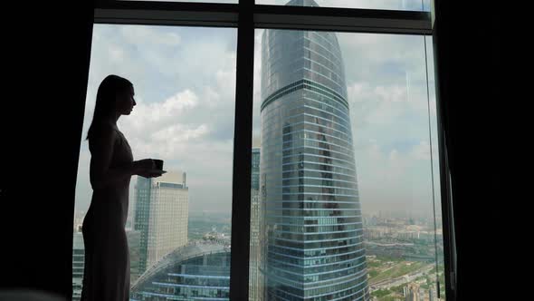 Woman in a Nightgown Approaches a Panoramic Window in a Skyscraper