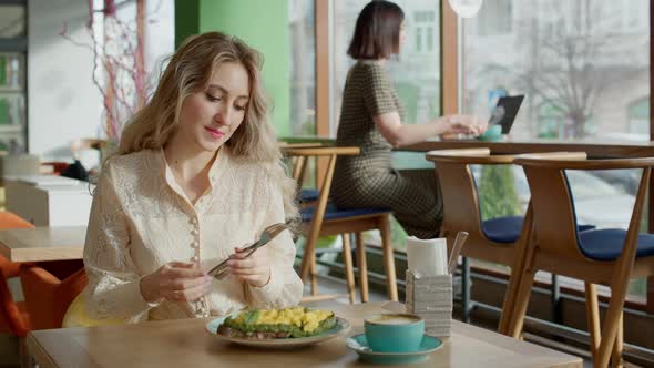 Relaxed Slim Caucasian Woman Eating Delicatessen in Cafe with Blurred Businesswoman Typing on Laptop