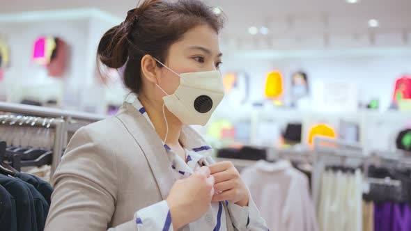 asian female shopper wearing facial virus protective mask choosing clothes at the store