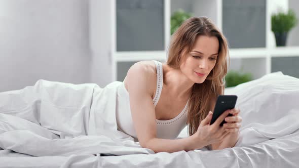 Smiling Young Woman Lying on Bed Relaxing Chatting Using Smartphone