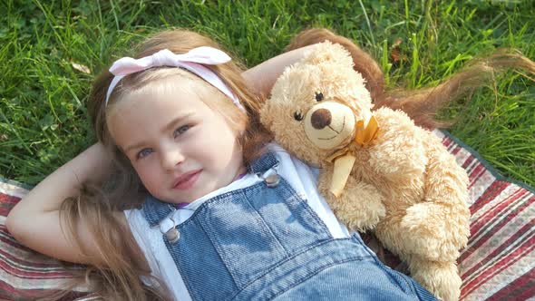 Pretty little child girl laying down with her teddy bear toy on blanket on green grass in summer