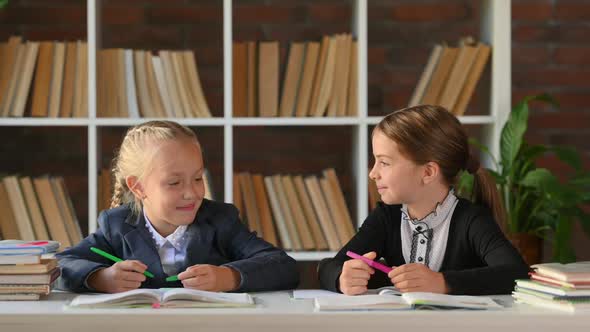 cute little schoolgirls sitting at table desk at library or home chatting and smiling