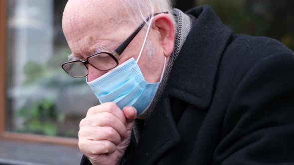 A Senior Man on the Street Coughs in a Protective Mask with Symptoms of Covid19