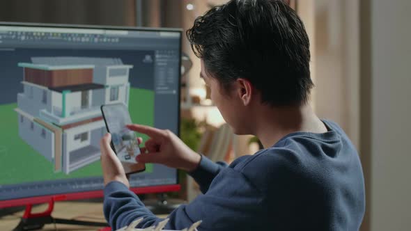 Asian Male Engineer Using Smartphone While Designing House On A Desktop At Home